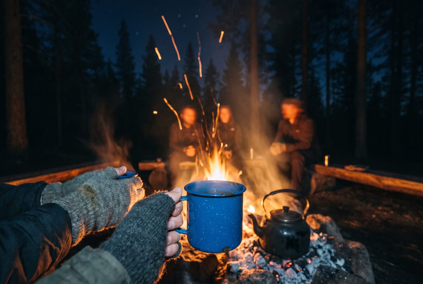 A campfire at night from where you sit: your hands in fingerless wool gloves cradling an enamel mug, sparks rising past a kettle on the coals, and friends as warm blurred figures on the log across the fire.